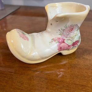 May include: A ceramic boot-shaped vase in cream with pink and red rose floral designs. The vase has a rounded toe and a flared top, and is sitting on a wooden surface.