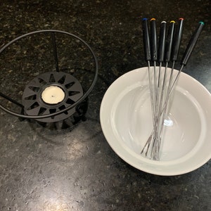 以下が含まれることがあります： 白いセラミック製のフォンデュ鍋と、黒い金属製のスタンド、カラフルなディッピングフォーク6本。スタンドは星形のデザインの円形ベースで、中央に小さなキャンドルホルダーがあります。