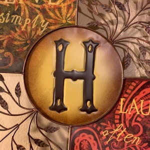 May include: A metal wall decor with a brown, yellow, and red color scheme. The center features a large letter "H" in brown with a yellow background. The surrounding sections feature the phrases "Live simply" and "Laugh often" in a decorative font.