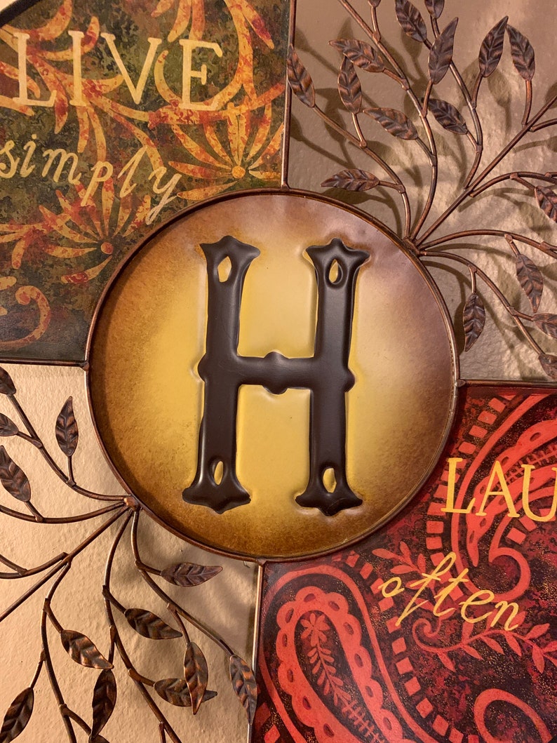 May include: A metal wall decor with a brown, yellow, and red color scheme. The center features a large letter "H" in brown with a yellow background. The surrounding sections feature the phrases "Live simply" and "Laugh often" in a decorative font.