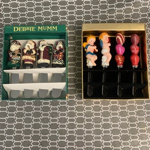 May include: Two sets of decorative cheese knives. One set features four Santa Claus figurines with red, green, and white accents. The other set features four cherub figurines with blue, red, white, and gold accents. The packaging for both sets includes the text "Debbie Mumm".