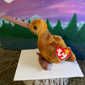 May include: A brown and tan stuffed kiwi bird with a long beak. The bird is sitting on a white surface with a brown tree stump underneath. The bird has a red Ty Beanie Baby tag attached to its wing.