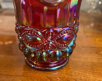 May include: A small, red iridescent carnival glass creamer featuring a scalloped rim and vertical fluting. The body is decorated with a band of embossed oval shapes, swirling patterns, and a row of raised circular beads around the base.