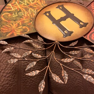May include: A metal wall decor featuring a tree with leaves and a circular plaque with the letter "H" in the center. The plaque has a yellow background and the letter is brown. The words "Live Simply" are written on one of the tree branches.