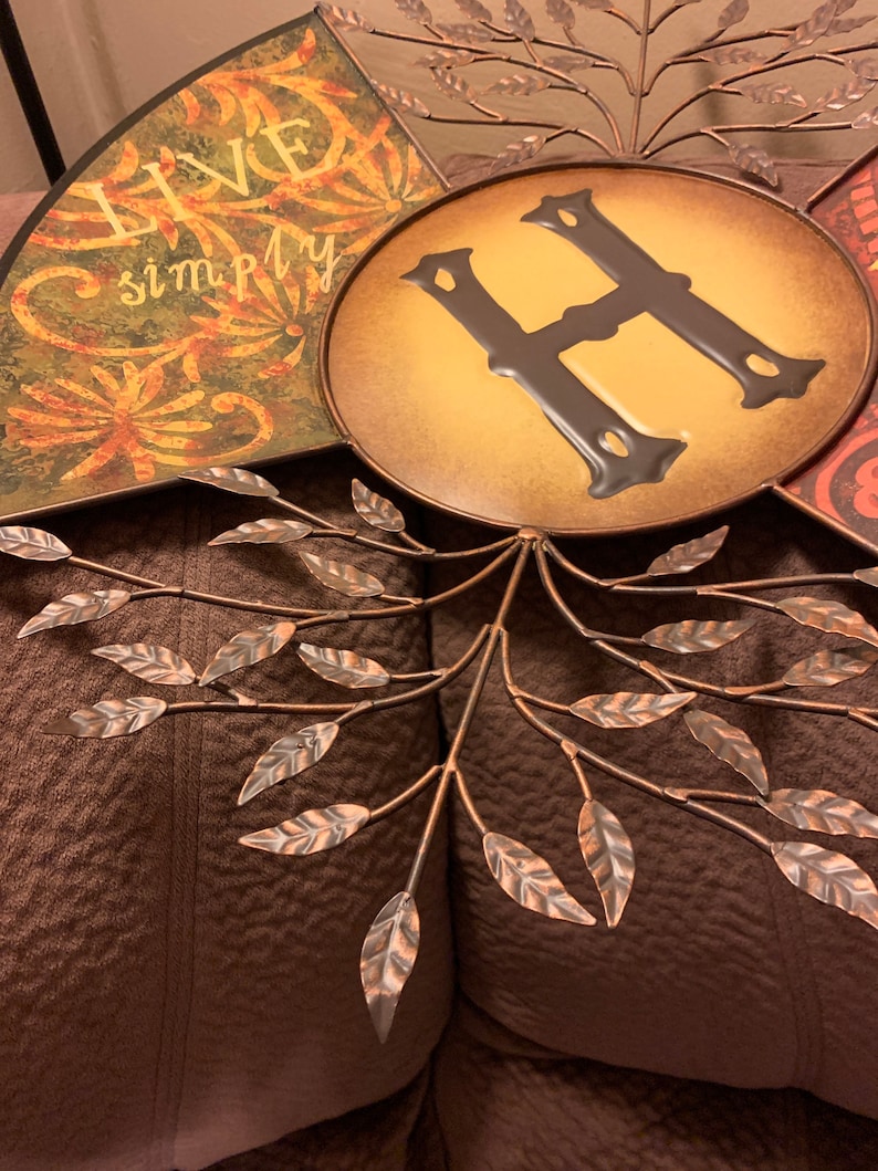 May include: A metal wall decor featuring a tree with leaves and a circular plaque with the letter "H" in the center. The plaque has a yellow background and the letter is brown. The words "Live Simply" are written on one of the tree branches.