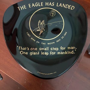 Puede incluir: Plato triangular negro con letras y diseño dorados que conmemoran el alunizaje del Apolo 11. El texto incluye "THE EAGLE HAS LANDED", "FIRST MAN ON THE MOON JULY 20, 1969" y una cita famosa. El plato presenta un águila y una representación de la luna.