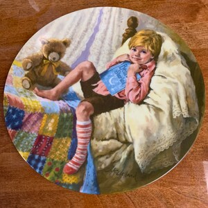 Könnte beinhalten: Ein Porzellanteller mit einem Gemälde eines jungen Jungen im Bett, der ein Buch mit dem Titel "Bedtime Stories" liest. Er trägt ein rosa Hemd, eine braune Hose und gestreifte Socken. Ein Teddybär mit einer gelben Schleife sitzt neben ihm. Das Bett ist mit einer bunten Patchwork-Decke bedeckt.