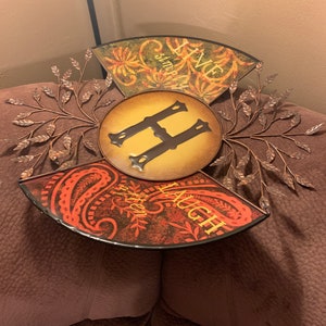 May include: A metal wall decor with a circular design featuring the letter "H" in the center. The three sections of the design are decorated with floral patterns and the words "Live Simply Laugh Often".