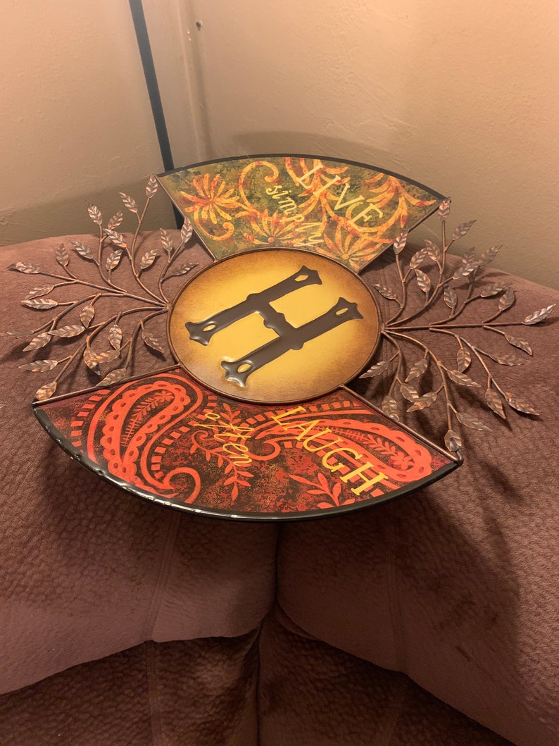 May include: A metal wall decor with a circular design featuring the letter "H" in the center. The three sections of the design are decorated with floral patterns and the words "Live Simply Laugh Often".