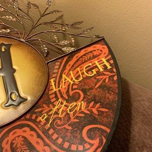 May include: A metal wall decor with a paisley pattern and the words "Laugh often" in gold lettering. The decor is in shades of red, brown, and gold.