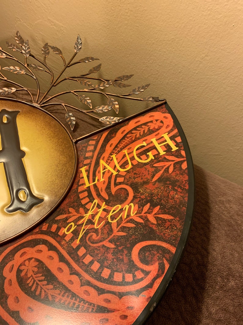 May include: A metal wall decor with a paisley pattern and the words "Laugh often" in gold lettering. The decor is in shades of red, brown, and gold.