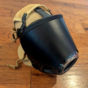 May include: A black plastic muzzle with a smooth, curved front panel and a clear circular opening at the tip. The device features a tan mesh fabric head strap system with adjustable black nylon webbing and plastic buckles for secure attachment.