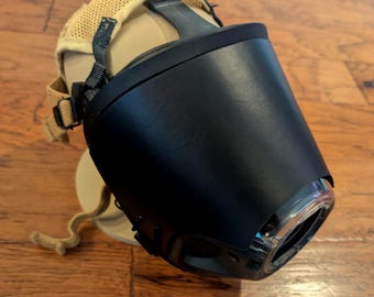 May include: A black plastic muzzle with a smooth, curved front panel and a clear circular opening at the tip. The device features a tan mesh fabric head strap system with adjustable black nylon webbing and plastic buckles for secure attachment.