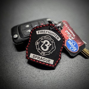 May include: Black leather firefighter keychain with red stitching. The keychain features a white firefighter helmet with the number 73 and the text "Firefighter" and "FD Sprague".