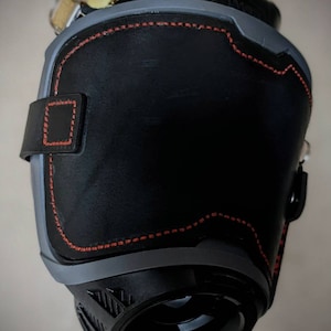May include: A black leather knee pad with red stitching and a gray plastic shell. The pad has a strap with a buckle.