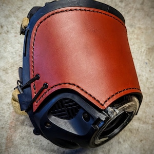 May include: A close-up of a black camera lens with a brown leather cover. The leather cover has stitching around the edges and a flap that covers the lens.