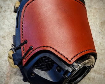 May include: A close-up of a black camera lens with a brown leather cover. The leather cover has stitching around the edges and a flap that covers the lens.