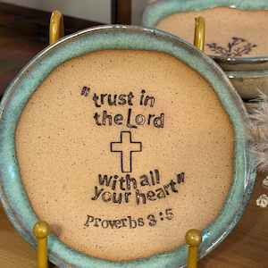 May include: A round ceramic plate with a teal rim and a tan center, held by a gold stand. The plate features the engraved text "trust in the Lord with all Your heart" and "Proverbs 3:5" along with a cross symbol.