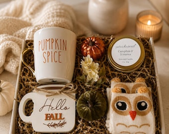 May include: A gift box containing a white mug with orange Pumpkin Spice text and a gold handle, a white wooden sign with Hello Fall text, a small metallic candle tin, a pair of owl-patterned socks, and two small decorative pumpkins.