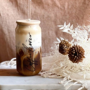 May include: A glass jar with a wooden lid filled with iced coffee. The jar has the words "Coffee Lover" written on it in black script. A clear glass straw is in the jar. The jar is sitting on a white marble surface.