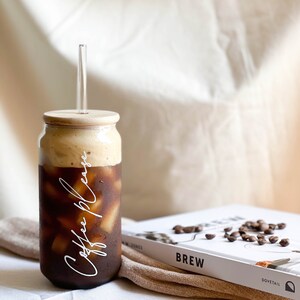 May include: A glass jar with a wooden lid and a clear straw filled with iced coffee. The jar has the text "Coffee Please" written on it in white. The jar is sitting on a white surface next to a book with the word "Brew" on the cover.