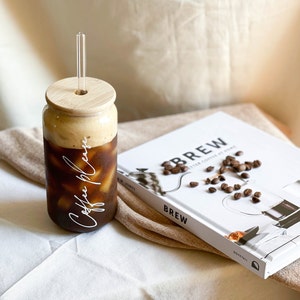 May include: A glass jar with a bamboo lid and a glass straw filled with iced coffee. The jar has the words "Coffee Please" written on it in white cursive. The jar is sitting on a white surface next to a book titled "Brew: Better Coffee at Home".