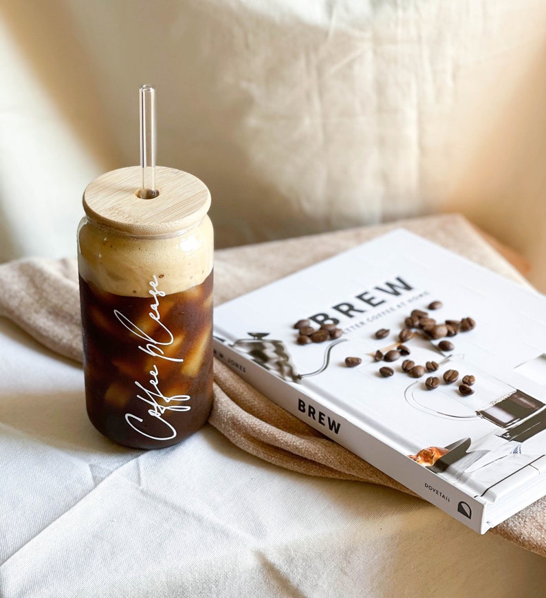May include: A glass jar with a bamboo lid and a glass straw filled with iced coffee. The jar has the words "Coffee Please" written on it in white cursive. The jar is sitting on a white surface next to a book titled "Brew: Better Coffee at Home".