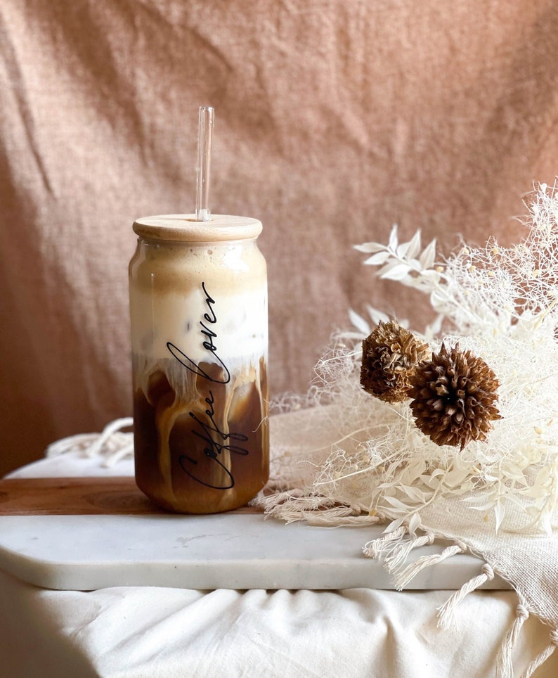 May include: A glass jar with a wooden lid filled with iced coffee. The jar has the words "Coffee Lover" written on it in black script. A clear glass straw is in the jar. The jar is sitting on a white marble surface.