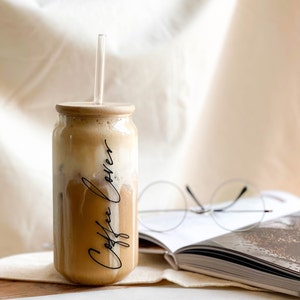 May include: A glass jar with a wooden lid and a straw filled with a light brown beverage. The jar has the words "Coffee Lover" written on it in black script. The jar is sitting on a white surface next to a book and glasses.