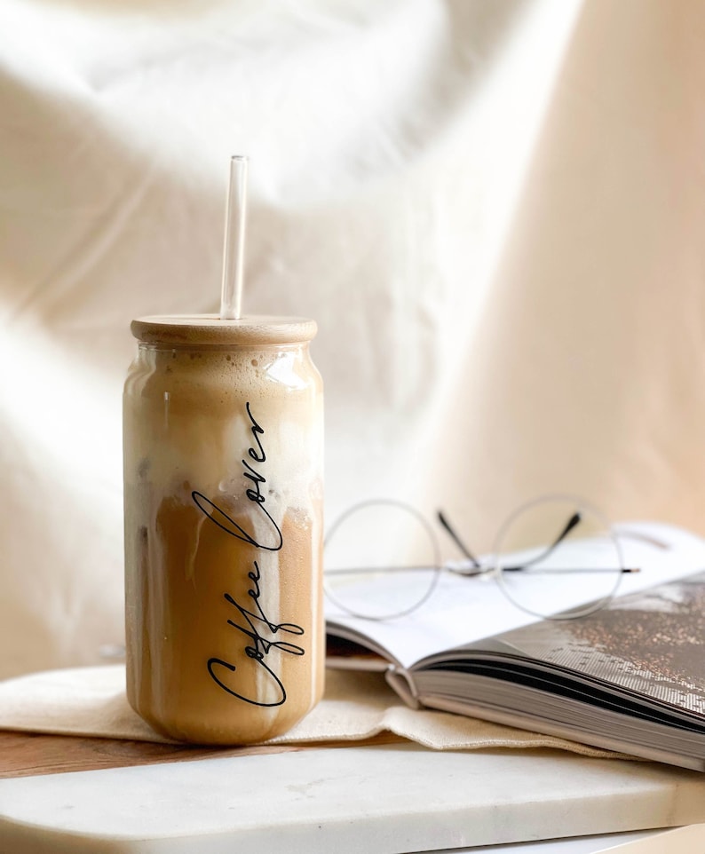 May include: A glass jar with a wooden lid and a straw filled with a light brown beverage. The jar has the words "Coffee Lover" written on it in black script. The jar is sitting on a white surface next to a book and glasses.