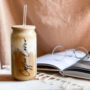May include: A glass can with a wooden lid filled with iced coffee and a glass straw. The can has the words "Coffee Lovers" written on it in black script. The can is sitting on a white marble surface next to a book and glasses.