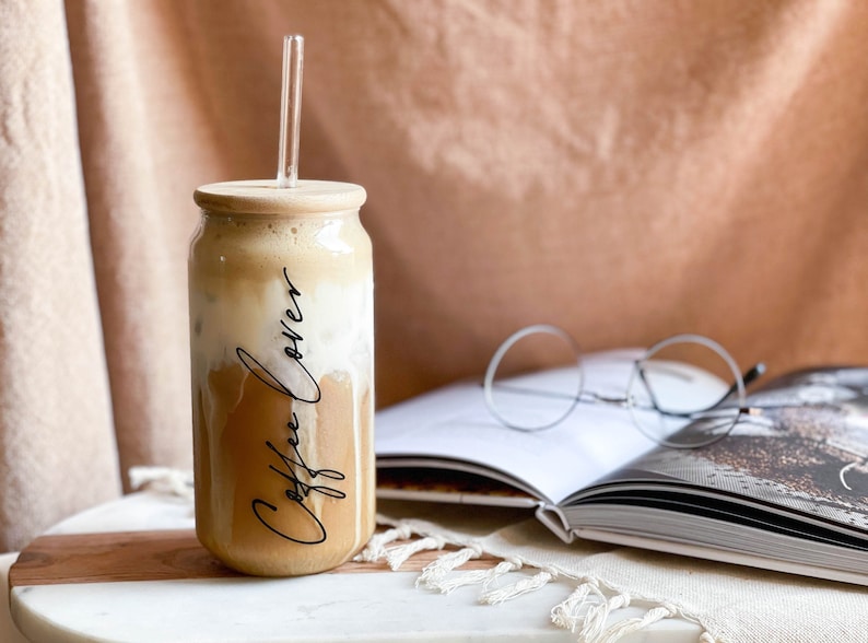 May include: A glass can with a wooden lid filled with iced coffee and a glass straw. The can has the words "Coffee Lovers" written on it in black script. The can is sitting on a white marble surface next to a book and glasses.