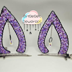 May include: A pair of purple beaded cat ear hair clips. The ears are made of wire and covered in various shades of purple beads. Each ear has a dangling chain with a crystal pendant. The clips are silver.