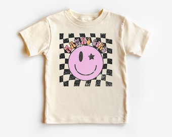 May include: A cream colored t-shirt with a pink smiley face with a star over one eye. The smiley face is surrounded by a black and white checkered pattern. The text "Mamas Girl" is written in a rainbow font above the smiley face.