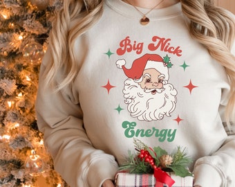 Könnte beinhalten: Ein beiger Pullover mit einer Retro-Grafik von Weihnachtsmann mit dem Text "Big Nick Energy" in Grün und Rot.
