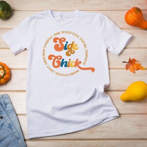 K&ouml;nnte beinhalten: Wei&szlig;es T-Shirt mit einem Grafikdesign, das "Side Chick" in einer Retro-Schriftart sagt. Die Grafik ist von einem Kreis aus Text umgeben, der verschiedene Thanksgiving-Beilagen auflistet.
