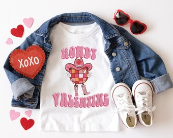 May include: A white t-shirt with a pink graphic of a disco ball wearing a cowboy hat and the text "Howdy Valentine". The shirt is paired with a denim jacket, red heart-shaped sunglasses, and white sneakers.