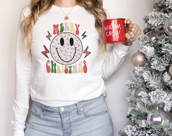 May include: White long-sleeved shirt with a pink, green, yellow, and red "Merry Christmas" graphic featuring a smiley face with a leopard print pattern. The shirt is worn by a person holding a red mug with the text "warm cozy" in white.
