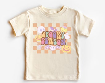May include: A cream colored t-shirt with a graphic of a checkered background with a variety of Halloween themed images including pumpkins, a ghost, a bat, a spider, a witch's hat, and the text "Spooky Season" in a retro font.