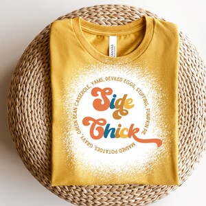 K&ouml;nnte beinhalten: Ein gelbes T-Shirt mit einer wei&szlig;en Grafik, auf der "Side Chick" in einer Retro-Schrift steht. Die Grafik ist von einem Kreis aus Text umgeben, der verschiedene Beilagen zum Thanksgiving-Essen auflistet.
