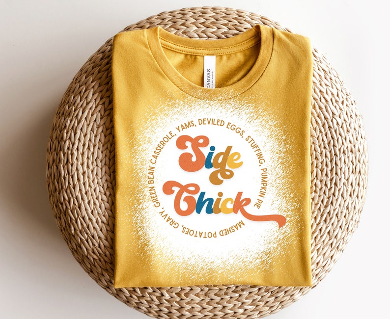 K&ouml;nnte beinhalten: Ein gelbes T-Shirt mit einer wei&szlig;en Grafik, auf der "Side Chick" in einer Retro-Schrift steht. Die Grafik ist von einem Kreis aus Text umgeben, der verschiedene Beilagen zum Thanksgiving-Essen auflistet.