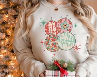 Könnte beinhalten: Ein cremefarbener Pullover mit einem Retro-Weihnachtsdesign mit drei Kugeln. Die Kugeln sind rot, pink und grün mit einem karierten Muster und dem Text "Ho Ho Ho" und "Frohe Weihnachten" in einer Retro-Schrift.