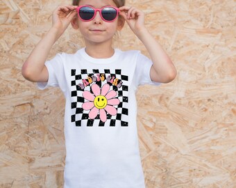 May include: White t-shirt with a black and white checkered pattern and a pink flower with a yellow smiley face. The text "Daddy's Girl" is written on the flower.