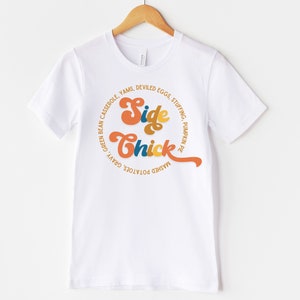 K&ouml;nnte beinhalten: Wei&szlig;es T-Shirt mit einem Grafikdesign, das "Side Chick" in einer Retro-Schriftart sagt. Die Grafik ist von einem Kreis aus Text umgeben, der verschiedene Thanksgiving-Beilagen auflistet.