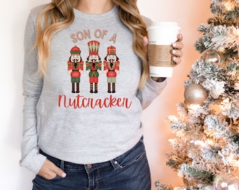 May include: A light gray long-sleeve shirt featuring a graphic of three nutcracker soldiers in red, green, and black. The text "SON OF A Nutcracker" is printed in red. The shirt is a festive holiday design.
