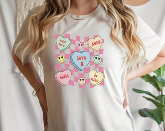 May include: White t-shirt with a pink and white checkered background and colorful heart-shaped candy graphics. The hearts have text on them that says "crazy for u", "xoxo", "love u", "cutie", and "be mine".