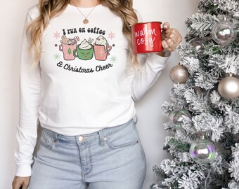May include: White long-sleeve shirt with a Christmas-themed design featuring three mugs of coffee and the text "I run on coffee & Christmas Cheer". The model is holding a red mug with the text "warm cozy".