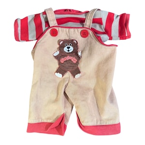Puede incluir: Un conjunto de peto de pana beige con una camiseta a rayas rojas y grises. El peto tiene ribetes rojos y botones rojos. Un aplique de oso de peluche marrón está en la parte delantera.