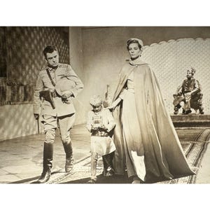 May include: Black and white photograph of a scene from a play or movie. A man in a military uniform walks beside a woman in a long cape. A child in a turban holds a book. A statue of a man sits in the background.