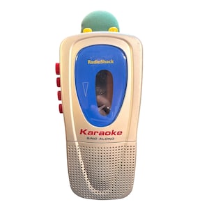 Op de afbeelding: Een vintage RadioShack Karaoke Sing-Along machine. Het apparaat is voornamelijk beige met een blauw middengedeelte en rode knoppen. De microfoon heeft een groene schuimrubberen hoes. De tekst "RadioShack" en "Karaoke SING-ALONG" zijn zichtbaar.
